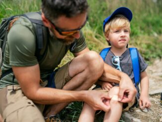 Father with small son hiking outdoors in summer nature, putting plaster on knee