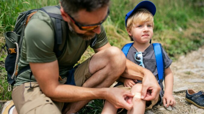 Father with small son hiking outdoors in summer nature, putting plaster on knee